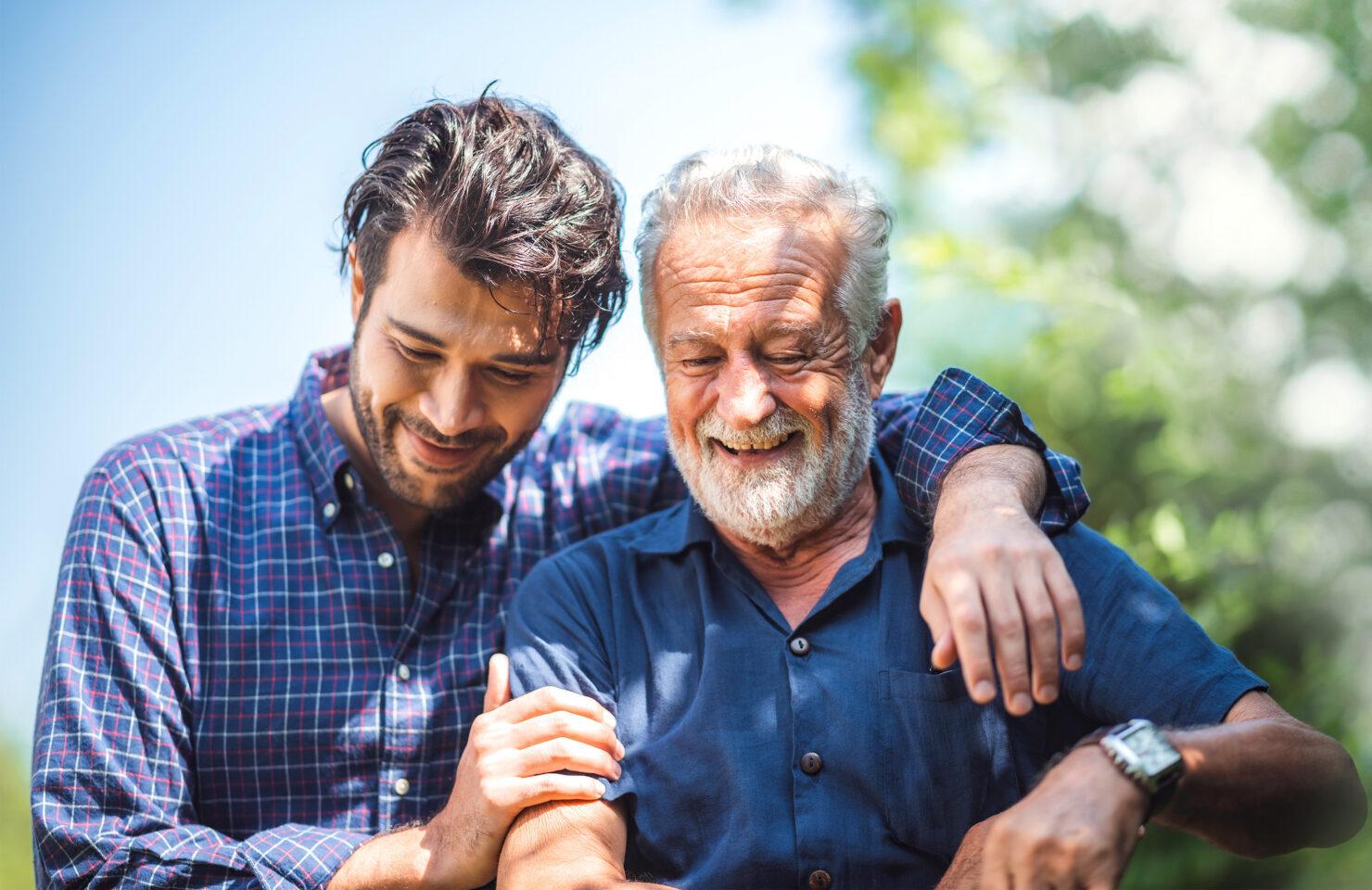 An adult son smiles with his arm around his father outdoors.