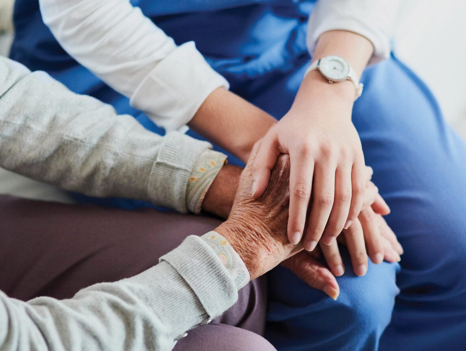 A compassionate nurse holds a senior patient’s hands. Their heads are out of frame.