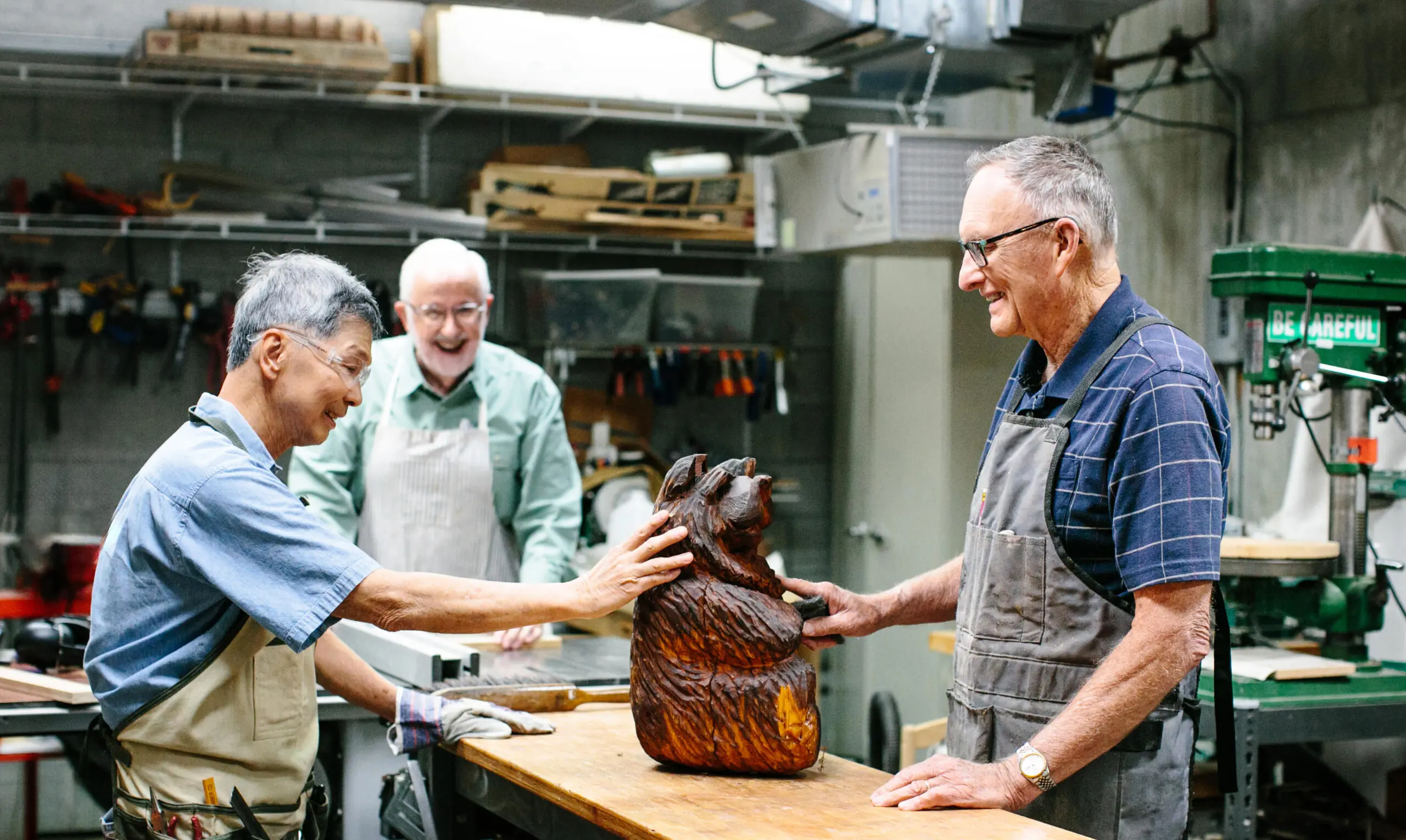 Three men stand in a woodshop and look proudly at an intricately-carved wooden creation.