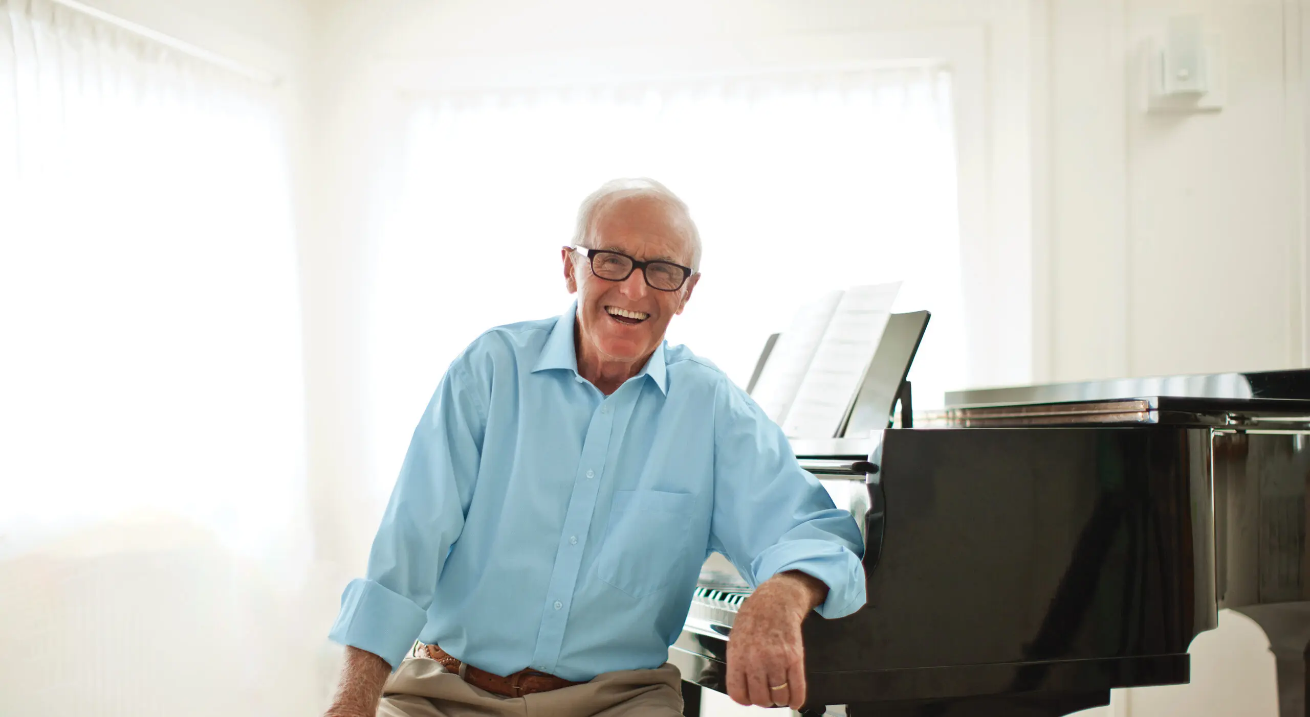 A smiling man sits on the bench of a baby grand piano.